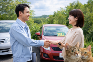 自社ローンや中古車買取の相談で笑顔で握手するお客様とスタッフ、車と猫が写る安心感のある店舗イメージ（岐阜美濃加茂）